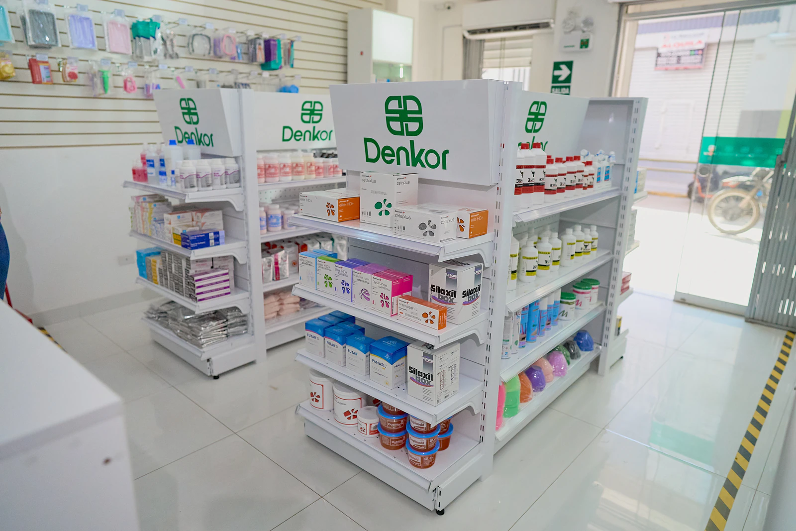 Interior del local, sin gente. Góndolas blancas con cabeceras de marca "Denkor" en verde, producto ordenado por categoría (cajas, frascos, siliconas…