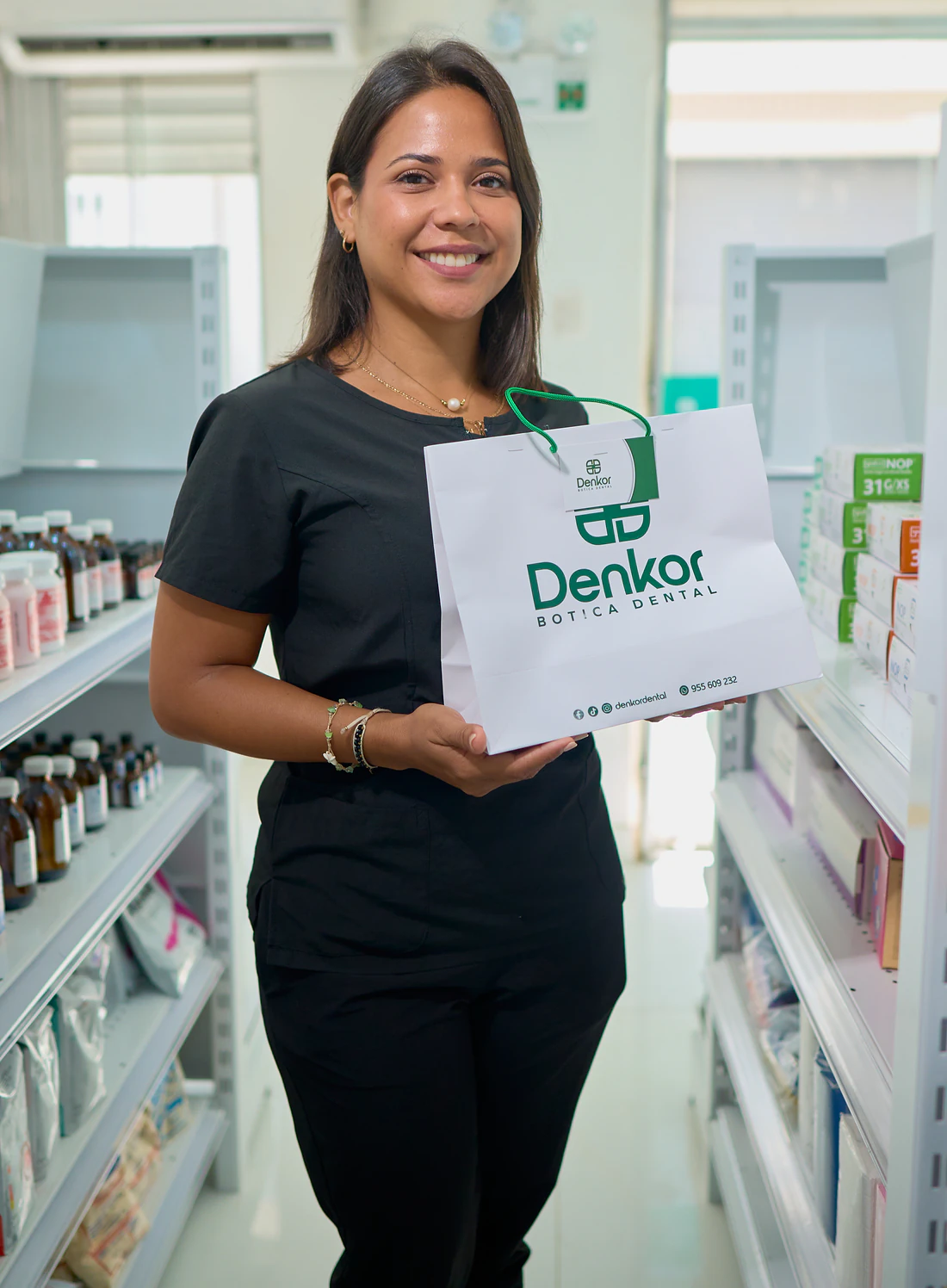 Colaboradora de uniforme negro, sonriendo de frente, sosteniendo la bolsa de papel de marca. Pasillo entre estanterías cargadas