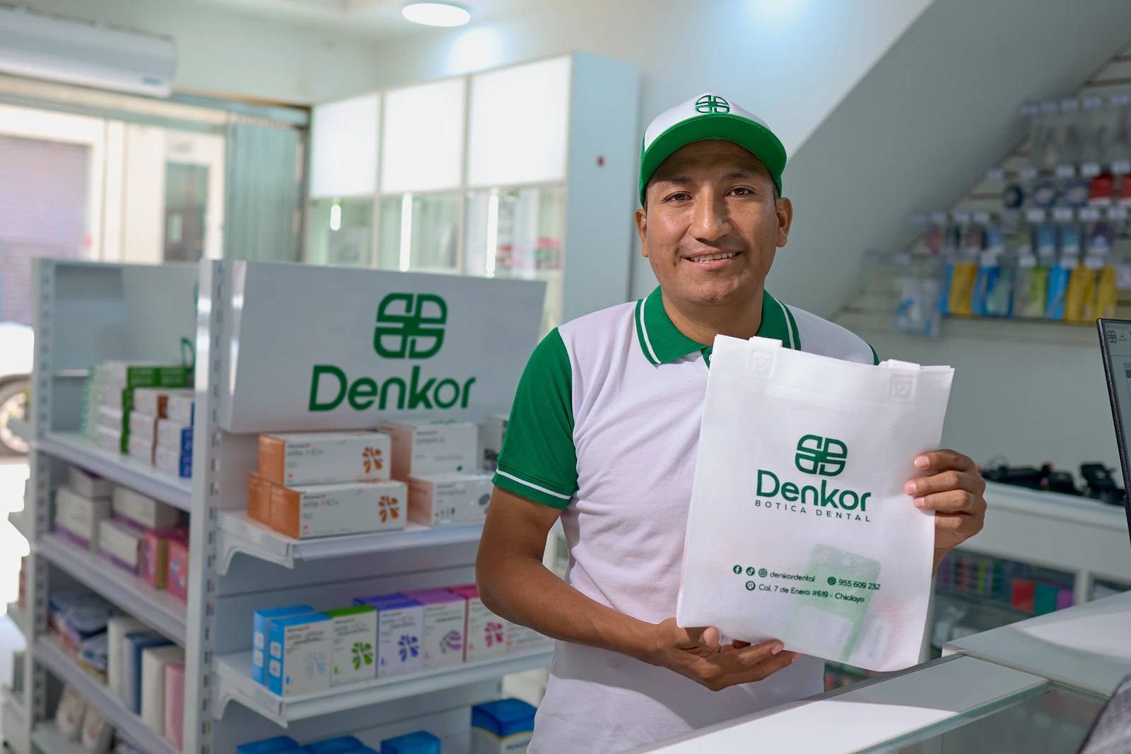 Colaborador con gorra y polo Denkor detrás del mostrador, con bolsa de marca en la que se lee la dirección: Cal. 7 de Enero #698 – Chiclayo. Góndola…