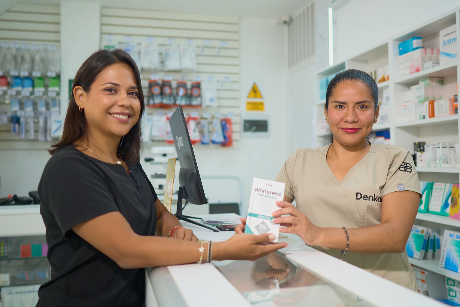 Dos personas en el mostrador: la clienta recibe una caja (Whiteness HP Maxx) de la colaboradora con uniforme Denkor. Estantería llena y panel de…